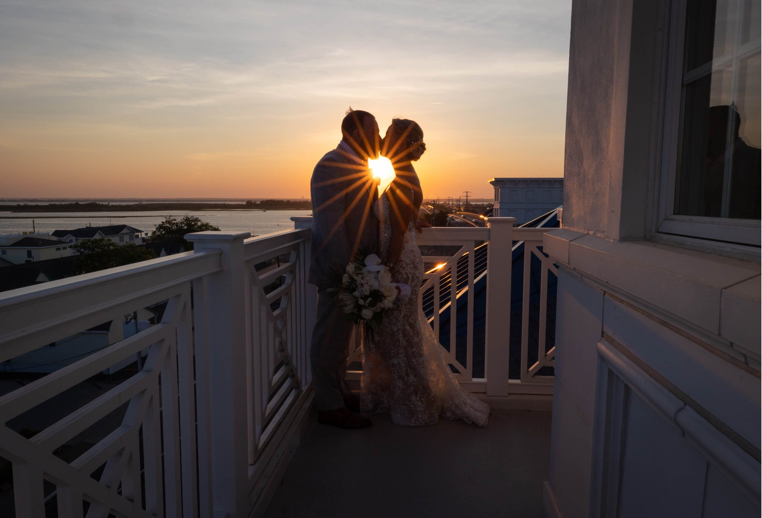 A couple stands on a balcony overlooking the water at sunset.