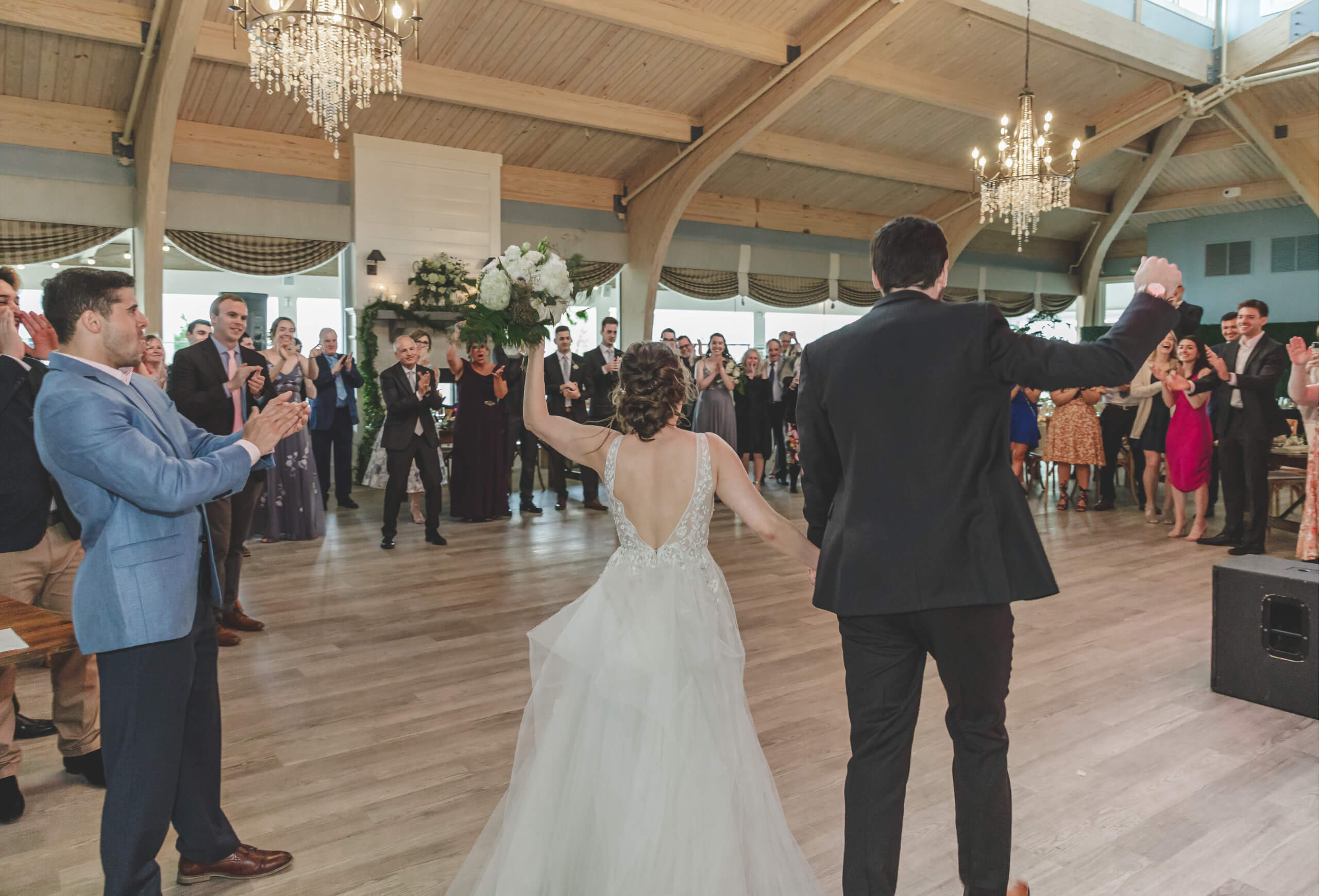 A large event space with a high ceiling, wood beams, and chandeliers, where a wedding party is celebrating.